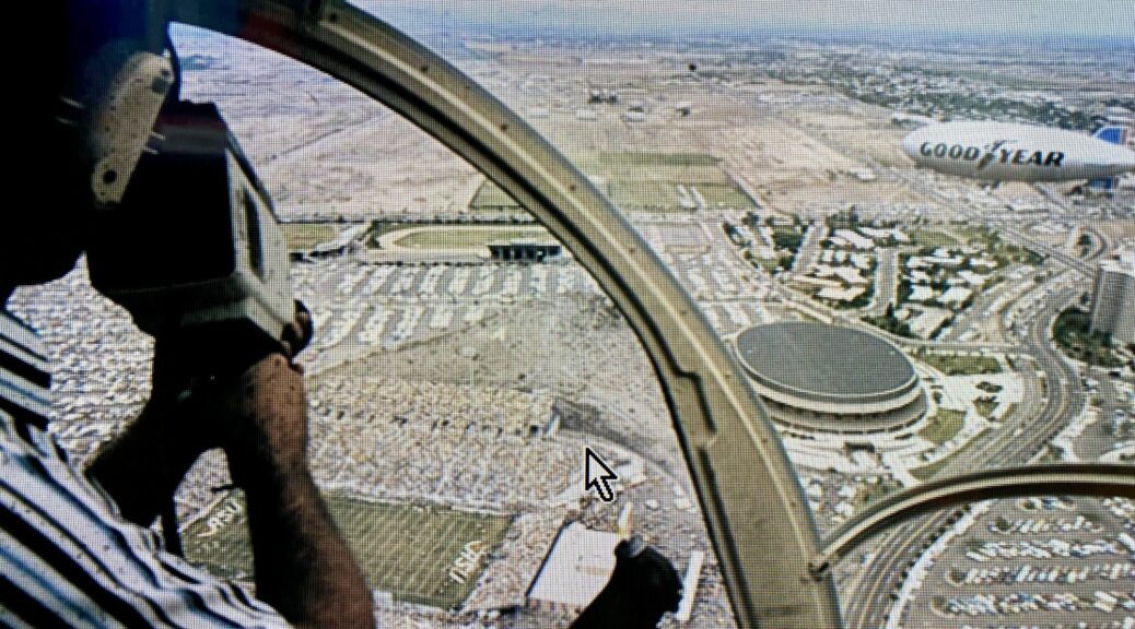 Pilot holding a handheld camera inside a helicopter cockpit, filming aerial footage.
