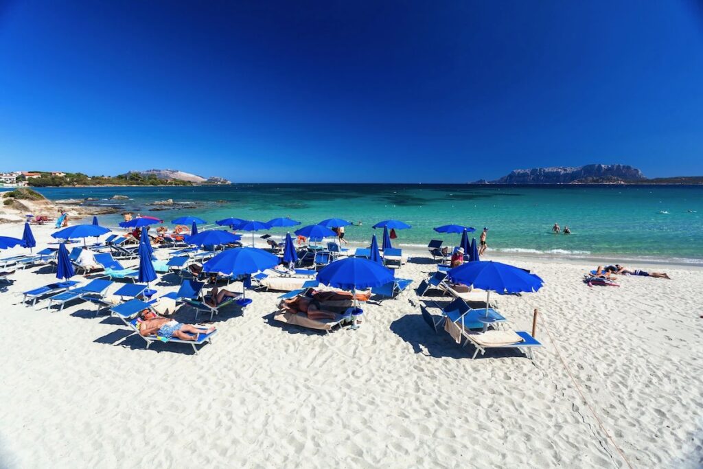 Beach at Hotel Stefania Boutique Hotel in Sardinia, Italy, with white sand, blue umbrellas and turquoise Mediterranean water.